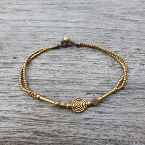 May include: A double-strand bracelet featuring small metallic gold-toned beads, two smooth cylindrical bars, two flat circular discs, and a central spiral charm. The bracelet is secured with a dark brown cord and a single spherical bead closure.