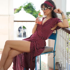 Puede incluir: Vestido burdeos con lunares blancos, mangas cortas y cuello en V. El vestido se combina con una diadema y gafas de sol a juego. La persona está sentada en una silla azul y bebiendo una bebida con pajita.