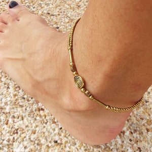 May include: A gold-toned anklet featuring a double-strand design composed of small spherical beads, smooth cylindrical spacers, and a central oval charm with a raised spiral pattern. The jewellery piece is crafted with metallic components.