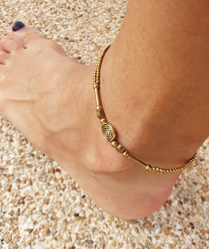 May include: A gold-toned anklet featuring a double-strand design composed of small spherical beads, smooth cylindrical spacers, and a central oval charm with a raised spiral pattern. The jewellery piece is crafted with metallic components.