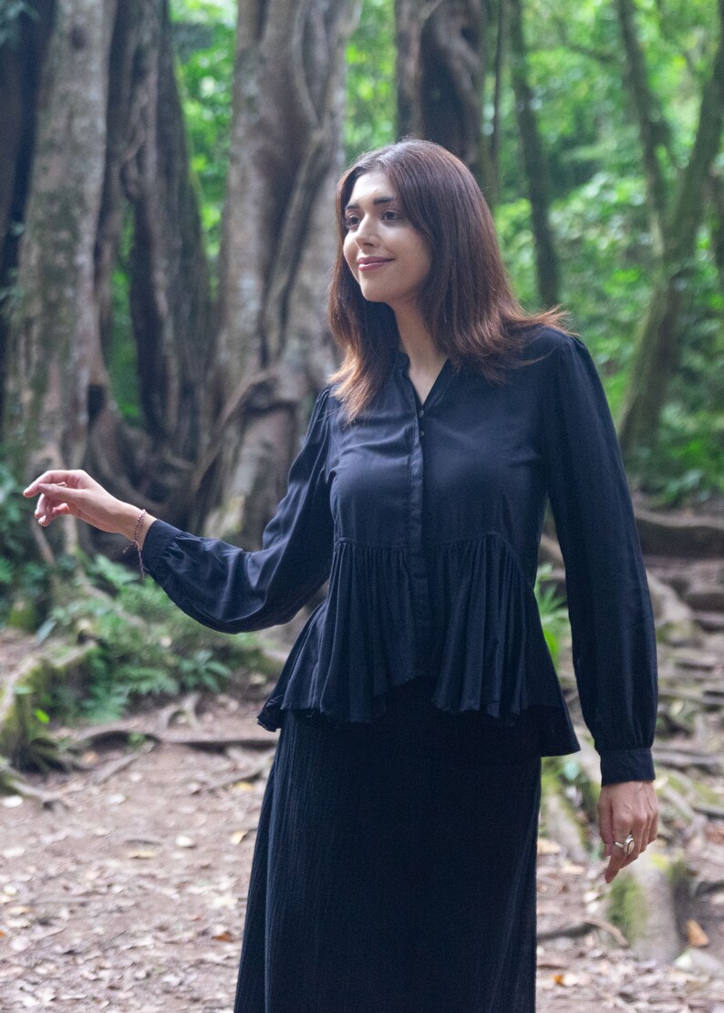 May include: A woman wearing a black long-sleeved blouse with a ruffled hem and a black skirt. She is standing in a forest setting.