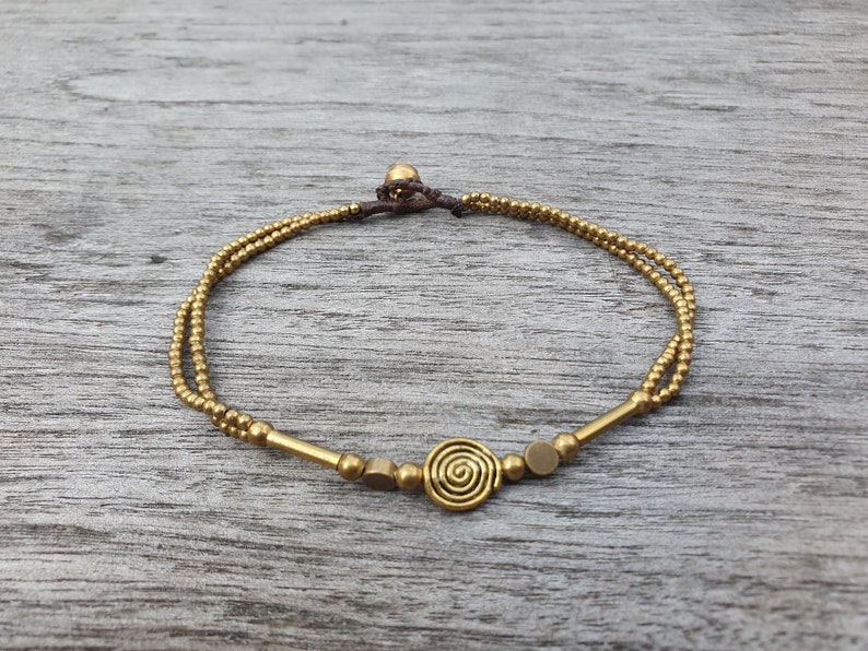 May include: A double-strand bracelet featuring small metallic gold-toned beads, two elongated cylindrical beads, two flat circular discs, and a central spiral-shaped charm. The bracelet is secured with a dark brown cord and a small spherical bead clasp.