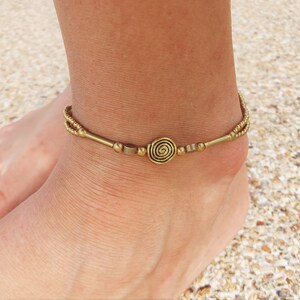 May include: A gold-toned metal anklet featuring a central spiral charm flanked by small cylindrical beads and spherical accents. The design includes a double-strand chain section on the sides, creating a layered appearance around the ankle.