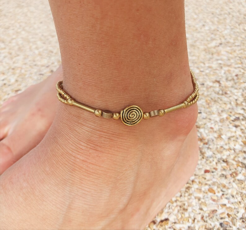 May include: A gold-toned metal anklet featuring a central spiral charm flanked by small cylindrical beads and spherical accents. The design includes a double-strand chain section on the sides, creating a layered appearance around the ankle.