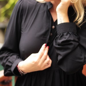 May include: A woman wearing a black long-sleeved blouse with a button-up front. The blouse has a ruffled hem and the woman is wearing a bracelet on her wrist.