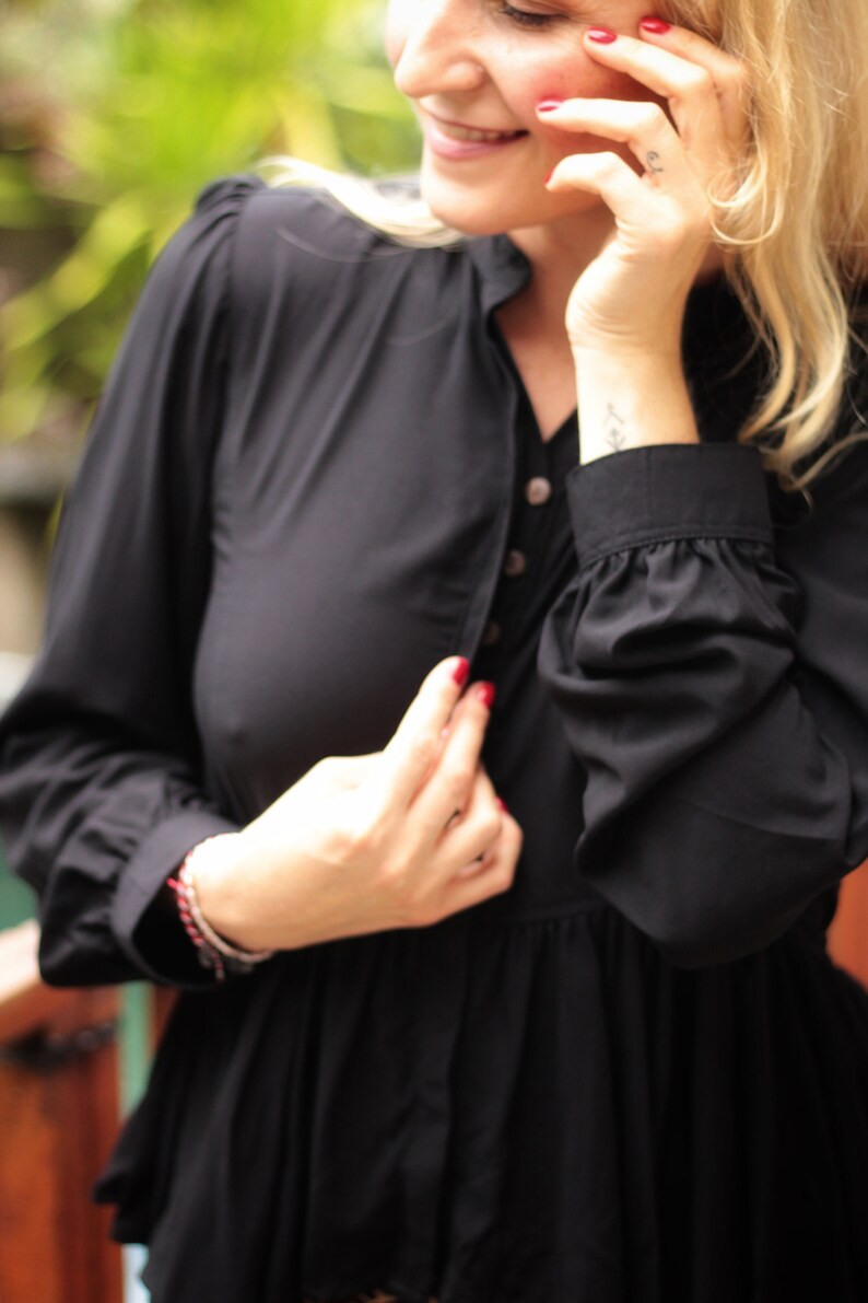 May include: A woman wearing a black long-sleeved blouse with a button-up front. The blouse has a ruffled hem and the woman is wearing a bracelet on her wrist.