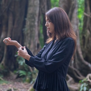 May include: A woman wearing a black long-sleeved blouse with a ruffled hem. The blouse has a high neckline and a button closure. She is standing in a forest setting.