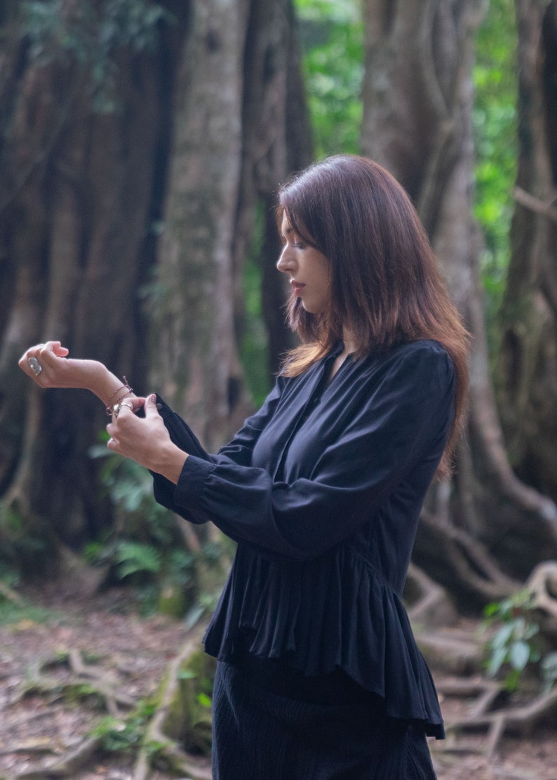 May include: A woman wearing a black long-sleeved blouse with a ruffled hem. The blouse has a high neckline and a button closure. She is standing in a forest setting.