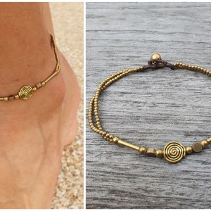 May include: A double-strand anklet featuring small metallic gold-toned beads, cylindrical spacers, and a central spiral charm. The piece is secured with a dark brown cord and a small bead closure, creating a layered look with metallic accents.