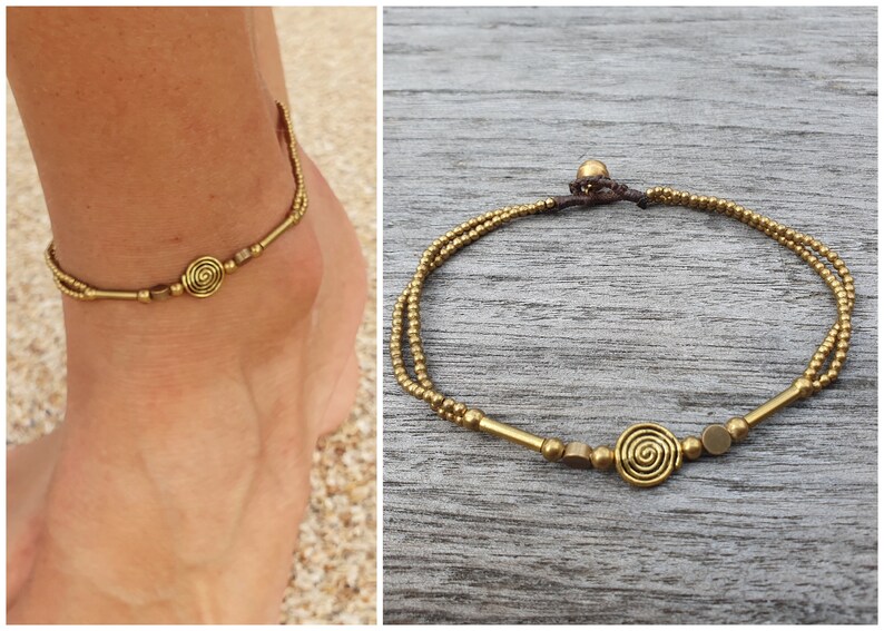 May include: A double-strand anklet featuring small metallic gold-toned beads, cylindrical spacers, and a central spiral charm. The piece is secured with a dark brown cord and a small bead closure, creating a layered look with metallic accents.
