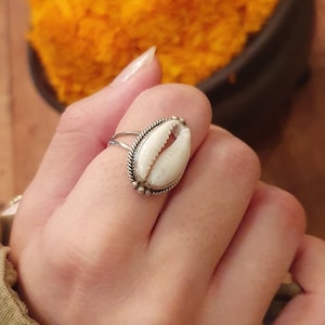 Könnte beinhalten: Ein silberner Ring mit einer weißen Kaurischnecke, die in einer dekorativen Fassung sitzt. Der Ring befindet sich an einem Finger.