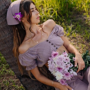 Könnte beinhalten: Eine Frau trägt ein lilafarbenes Top mit offener Schulter und ein lilafarbenes Bandana mit einer Blume. Sie hält einen Strauß rosa und weißer Blumen.