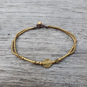 May include: A double-strand bracelet featuring small metallic gold-toned beads, two smooth cylindrical bars, two flat circular discs, and a central spiral charm. The bracelet is secured with a dark brown cord and a single spherical bead closure.