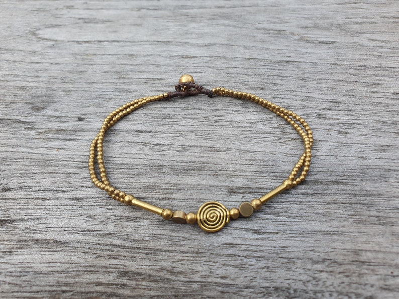 May include: A double-strand bracelet featuring small metallic gold-toned beads, two smooth cylindrical bars, two flat circular discs, and a central spiral charm. The bracelet is secured with a dark brown cord and a single spherical bead closure.