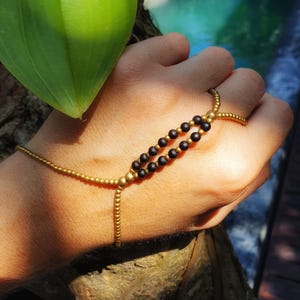 May include: A hand chain featuring a delicate gold-toned beaded bracelet connected to a ring by a central bridge. The bridge consists of two parallel rows of small matte black beads framed by gold-toned beads, creating a structured geometric design.