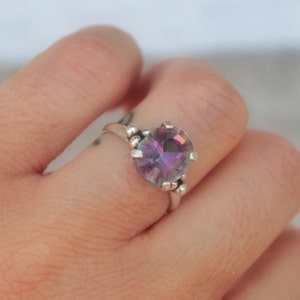 Op de afbeelding: Een zilveren ring met een grote, regenboogkleurige, paarse edelsteen die in een bezel-setting is gezet. De ring heeft een geperst bandje.