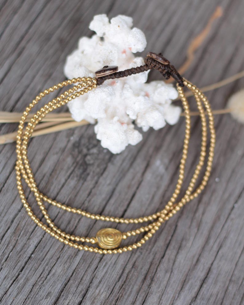 May include: A multi-strand bracelet featuring three rows of small metallic gold beads. The strands are connected by a dark brown cord with a knot closure. A single, larger, circular gold-toned bead with a spiral pattern is centred on one strand.