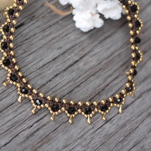 May include: A circular necklace featuring a dark brown woven cord base adorned with small metallic gold beads and faceted black beads. The design includes a repeating scalloped pattern with gold bead dangles hanging from the lower edge.