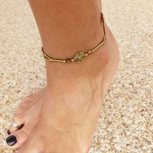 May include: A gold-toned anklet featuring a central spiral charm flanked by small cylindrical and spherical beads. The piece is constructed with a double-strand design, utilising thin metallic chains that connect to the decorative focal point.
