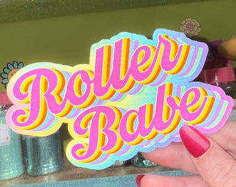 May include: A colorful sticker with the words "Roller Babe" in a retro font. The sticker has a layered design with pink, yellow, and blue hues. The background includes glitter jars and a shelf.