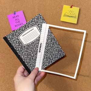 May include: A composition notebook with a black and white speckled cover, a white lined page, and a white square frame. A purple sticky note with the text "READ ALL THE BOOKS" is pinned to the background. A yellow sticky note with a book illustration is also pinned.