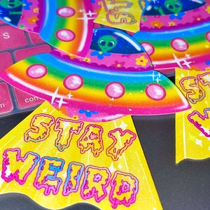 Peut inclure: Un ensemble de quatre autocollants color&eacute;s repr&eacute;sentant un vaisseau spatial arc-en-ciel sur fond rose avec le texte "Stay Weird".