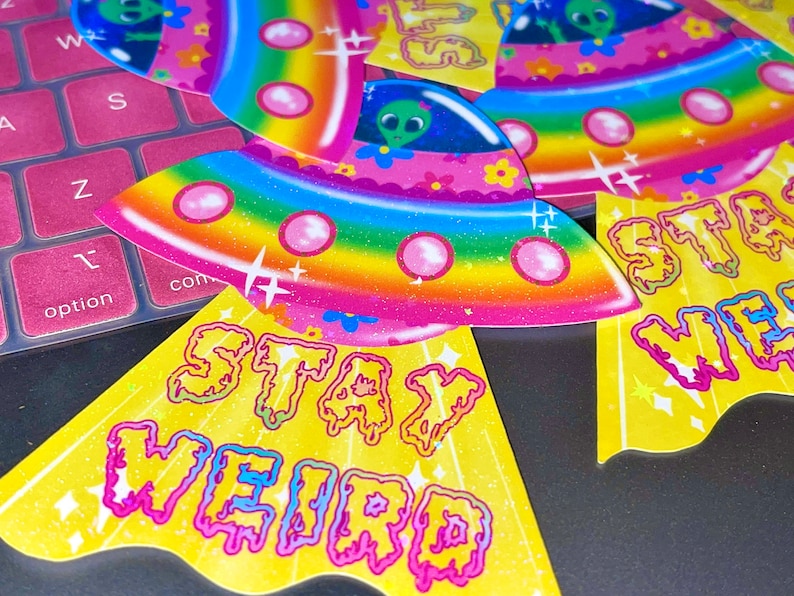 Peut inclure: Un ensemble de quatre autocollants color&eacute;s repr&eacute;sentant un vaisseau spatial arc-en-ciel sur fond rose avec le texte "Stay Weird".