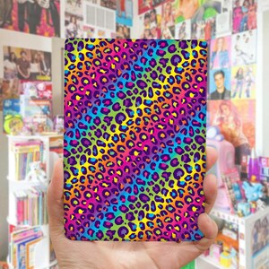 May include: A rectangular flat object featuring a vibrant rainbow-colored leopard print pattern. The design consists of dark purple and black animal spots layered over a gradient background that transitions through shades of yellow, pink, blue, and green.