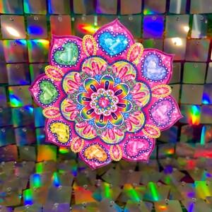 May include: A pink and rainbow mandala with hearts and jewels. The mandala is against a background of iridescent sequins.