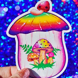 May include: A colorful mushroom sticker with a rainbow-colored top and a pink body. The sticker features three smaller mushrooms inside, with green leaves and a white outline. The background is a sparkly blue.