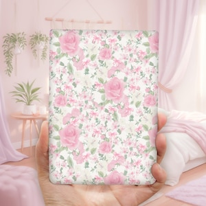 May include: A rectangular item with a floral pattern of pink roses and smaller pink flowers on a cream background. The item is held in a hand, with a soft pink background visible.