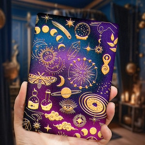 May include: A purple and blue notebook cover with a gold foil design featuring celestial and mystical symbols, including stars, moons, a butterfly, a crystal, and a sun.