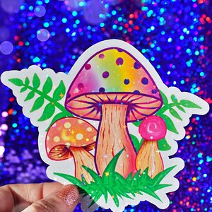 May include: A colorful sticker featuring three cartoon mushrooms with rainbow-colored caps and brown stems. The sticker has green grass and leaves around the mushrooms. The background is a sparkly blue.