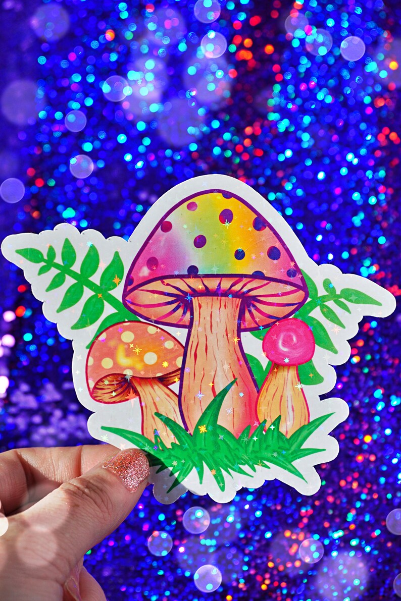 May include: A colorful sticker featuring three cartoon mushrooms with rainbow-colored caps and brown stems. The sticker has green grass and leaves around the mushrooms. The background is a sparkly blue.