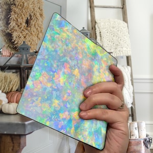 May include: A iridescent opal-like pattern on a square-shaped notebook cover. The cover has a shimmery, pearlescent finish with a variety of colors, including blue, green, pink, and yellow.
