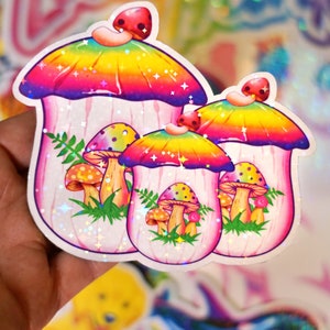 May include: A set of three mushroom stickers with a rainbow gradient cap and a pink stem. The stickers have a holographic, glittery finish. Smaller mushrooms and green foliage are also featured in the design.