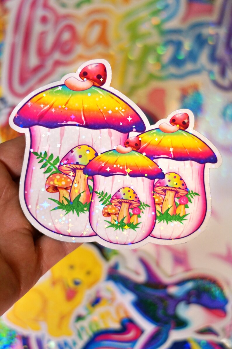 May include: A set of three mushroom stickers with a rainbow gradient cap and a pink stem. The stickers have a holographic, glittery finish. Smaller mushrooms and green foliage are also featured in the design.