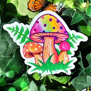 May include: A colorful sticker featuring three cartoon mushrooms with rainbow caps and orange stems, set against green grass. A butterfly with orange and black wings is perched above the mushrooms, with more butterflies in the background. The sticker is surrounded by green leaves.