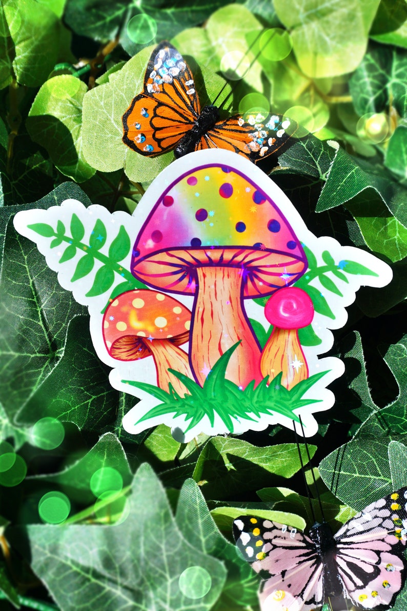 May include: A colorful sticker featuring three cartoon mushrooms with rainbow caps and orange stems, set against green grass. A butterfly with orange and black wings is perched above the mushrooms, with more butterflies in the background. The sticker is surrounded by green leaves.
