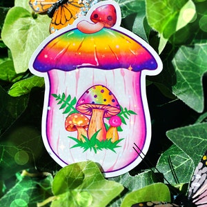 May include: A colorful sticker featuring a whimsical mushroom house design. The mushroom has a rainbow-colored top and houses three mushrooms inside. The sticker is surrounded by green leaves and butterflies.