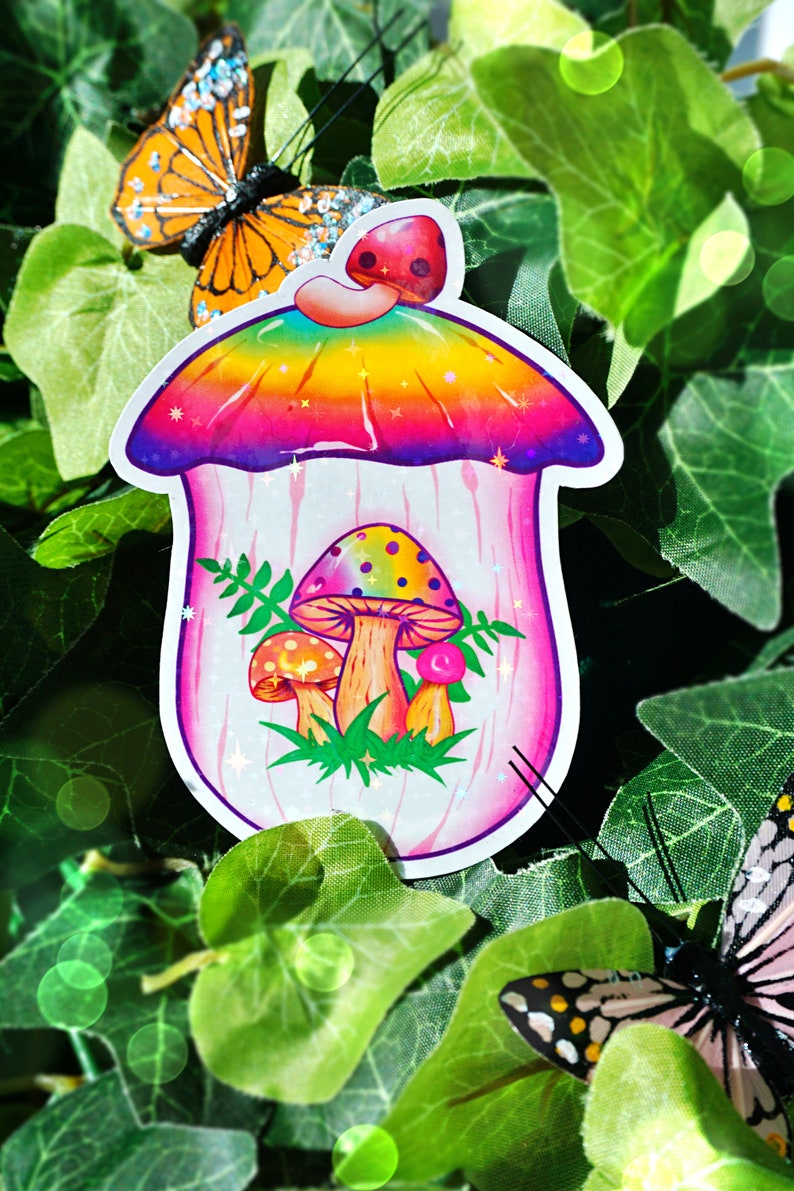 May include: A colorful sticker featuring a whimsical mushroom house design. The mushroom has a rainbow-colored top and houses three mushrooms inside. The sticker is surrounded by green leaves and butterflies.