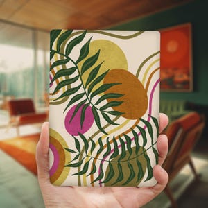 May include: A rectangular notebook with a cream-colored background featuring a botanical design. The design includes green leaves, brown and yellow circles, and pink and brown organic shapes. The notebook is held in a person's hand.