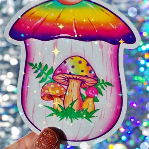 May include: A holographic sticker featuring a whimsical mushroom design. The mushroom has a rainbow-colored cap and a pink stem, with smaller mushrooms and green foliage inside. The sticker has a sparkly, iridescent finish.