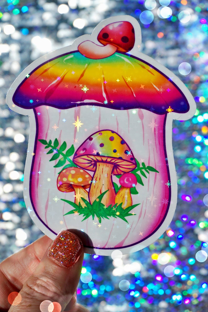 May include: A holographic sticker featuring a whimsical mushroom design. The mushroom has a rainbow-colored cap and a pink stem, with smaller mushrooms and green foliage inside. The sticker has a sparkly, iridescent finish.