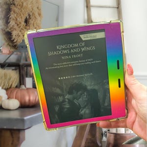 May include: A Kindle e-reader with a rainbow-colored frame. The screen displays the book cover for "Kingdom of Shadows and Wings" by Nina Frost. The cover art features a couple embracing. The e-reader is held in front of a blurred background.