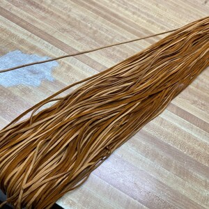 May include: A bundle of brown leather cord, approximately 1/4 inch wide, laid out on a wooden surface.
