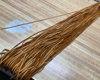 May include: A bundle of brown leather cord, approximately 1/4 inch wide, laid out on a wooden surface.