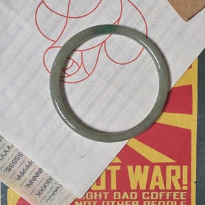 May include: A smooth, circular bangle bracelet with a translucent, pale green hue. The bangle has a polished, rounded surface and a consistent thickness throughout its entire circumference, presenting a simple and solid form without any added embellishments.