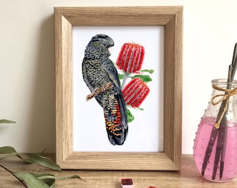 May include: A framed watercolor print featuring a black cockatoo perched on a branch with red flowers. The bird has detailed feather patterns in black, gray, and orange. The artwork is in a light wood frame, with watercolor paints and a vase in the background.