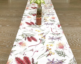 May include: White table runner with a floral pattern featuring red, yellow, and purple flowers. The runner is on a wooden table with a small potted plant in the center.